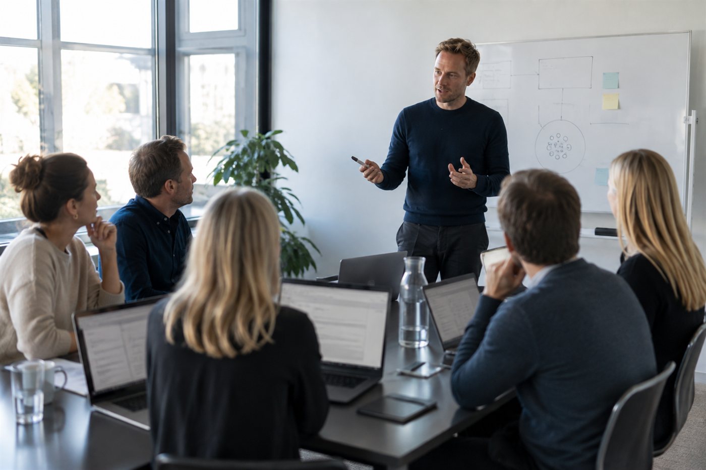 Rådgiver præsenterer en AI-arbejdsgang på et whiteboard for et team med bærbare computere i et mødelokale